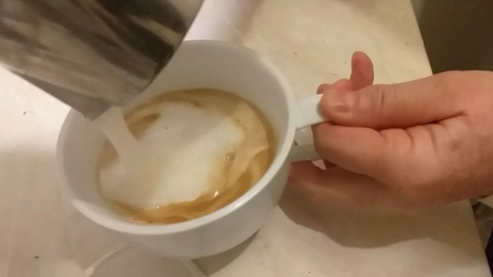 Eine Hand h&auml;lt eine Tasse, w&auml;hrend Hafermilch in den Kaffee gegossen wird, um einen cremigen Kaffee zu kreieren.