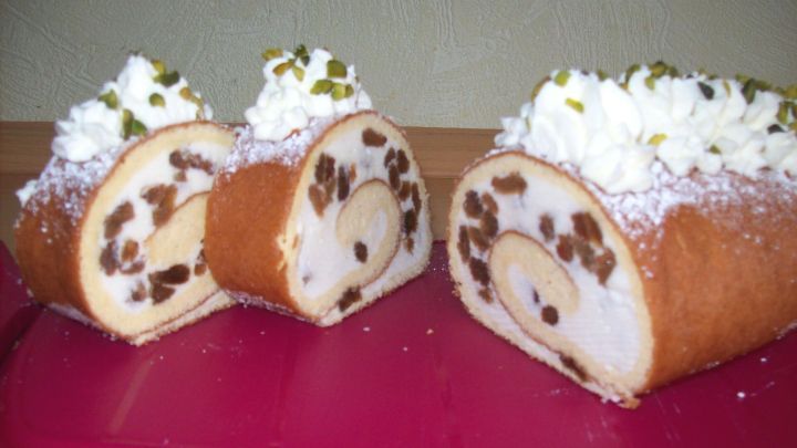 Eine Biskuit-Quarkrolle mit Rumrosinen, garniert mit Sahne und Pistazien, liegt auf einem pinken Teller.