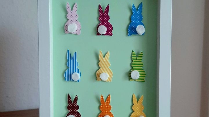 Ein Bilderrahmen zeigt neun bunte, gestickte Hasen auf einem gr&uuml;nen Hintergrund. Die Hasen sind in verschiedenen Farben und Mustern angeordnet.