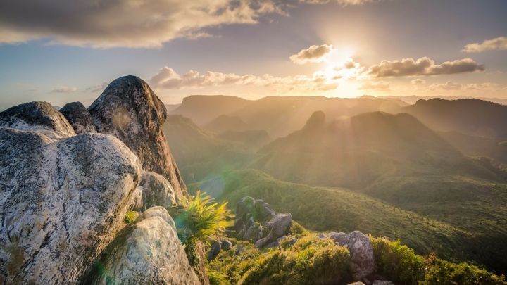 Ein majest&auml;tischer Felsen steht im Vordergrund, w&auml;hrend die Sonne hinter den Bergen aufgeht und Hoffnung in die Landschaft bringt.