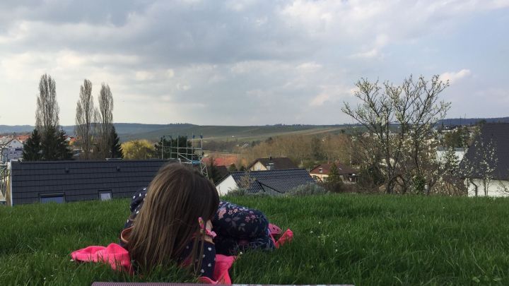 Ein Kind liegt entspannt auf einer Wiese und betrachtet die Landschaft unter einem bew&ouml;lkten Himmel.