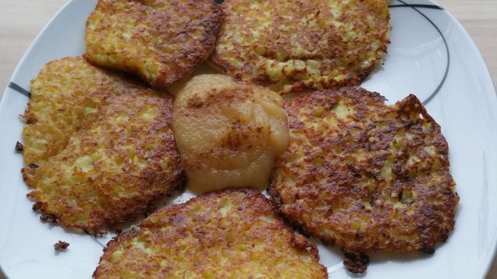 Sechs goldbraune Kartoffelpuffer liegen auf einem wei&szlig;en Teller, umgeben von einer hellen Holzoberfl&auml;che.