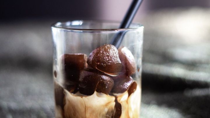 Ein Glas Eiskaffee mit Kaffeew&uuml;rfeln und einem schwarzen Strohhalm steht auf einem schlichten, dunklen Tisch.