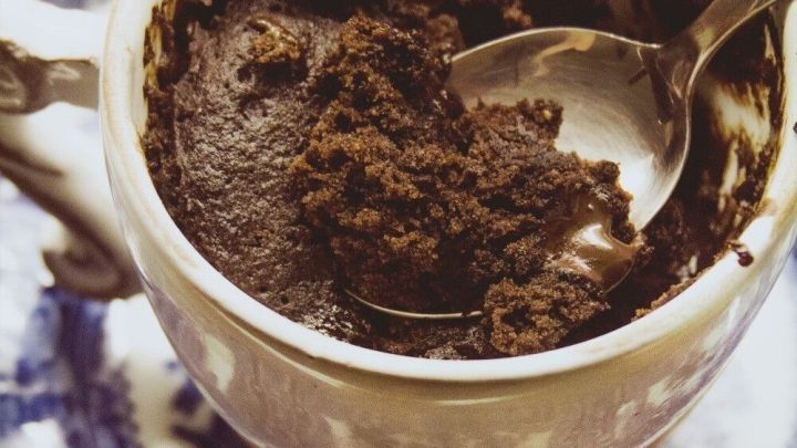 Ein Tassenkuchen aus Schokolade sitzt in einer verzierteren Tasse, w&auml;hrend ein L&ouml;ffel die weiche, feuchte Oberfl&auml;che durchdringt.