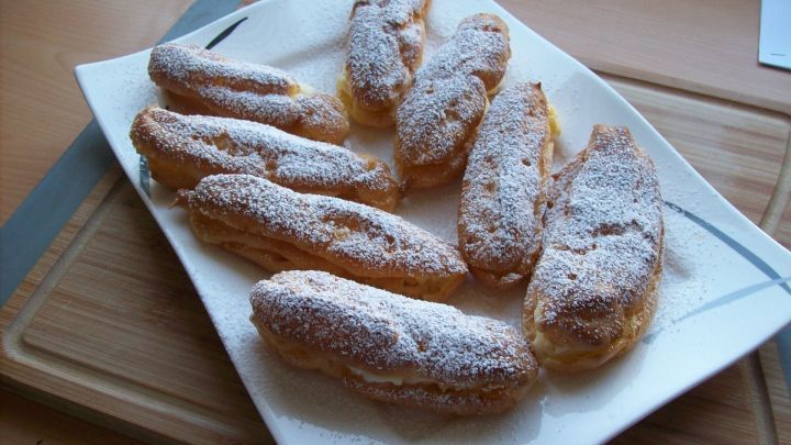 Sechs frisch gebackene Brandteiggeb&auml;cke liegen auf einem wei&szlig;en Teller und sind mit Puderzucker bestreut, auf Holzuntergrund.