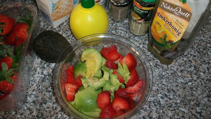 Eine Sch&uuml;ssel mit Erdbeer-Guacamole steht vor verschiedenen Zutaten wie Avocado, Zitronensaft und Gew&uuml;rzen auf einer Arbeitsplatte.
