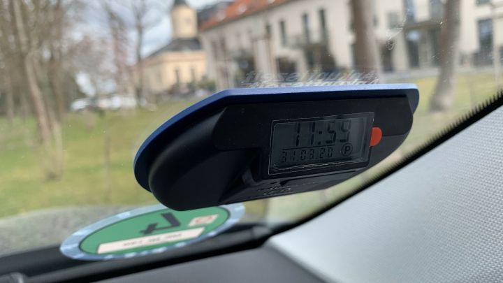 Eine elektronische Parkscheibe zeigt die Uhrzeit an und wird im Auto verwendet, um Parkzeiten zu regulieren, w&auml;hrend im Hintergrund ein Geb&auml;ude sichtbar ist.
