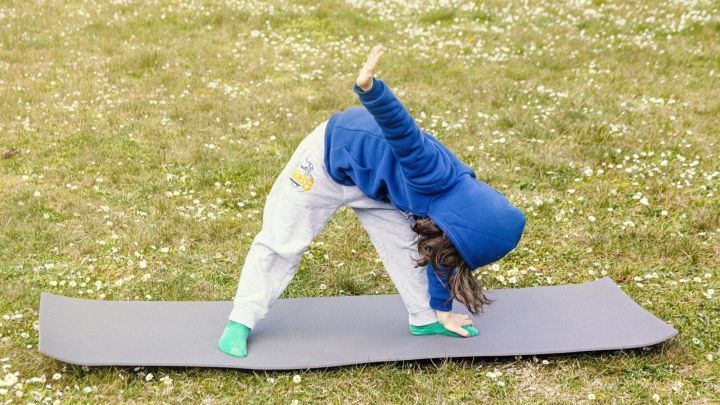 Ein Kind &uuml;bt eine Yoga-Position auf einer Matte in einer bl&uuml;henden Wiese, umfit mit einem blauen Hoodie und wei&szlig;en Hosen.