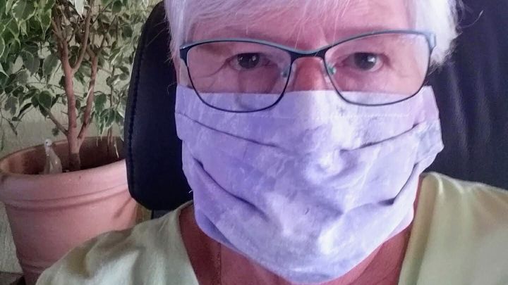 Eine Frau mit einer selbst gen&auml;hten Community-Maske sitzt entspannt vor einem gr&uuml;nen Pflanzenarrangement in ihrem Zuhause.