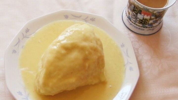 Auf einem Teller liegt eine gro&szlig;e, feuchte Dampfnudel in einer s&uuml;&szlig;en So&szlig;e, neben einer Tasse mit Kaffee.