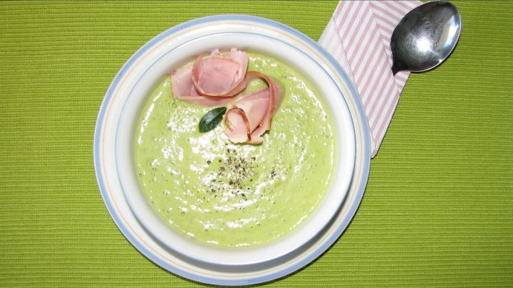 Eine cremige Erbsenrahmsuppe mit zarten Schinkenrosen und frischen Kr&auml;utern in einer wei&szlig;en Sch&uuml;ssel auf gr&uuml;nem Hintergrund.