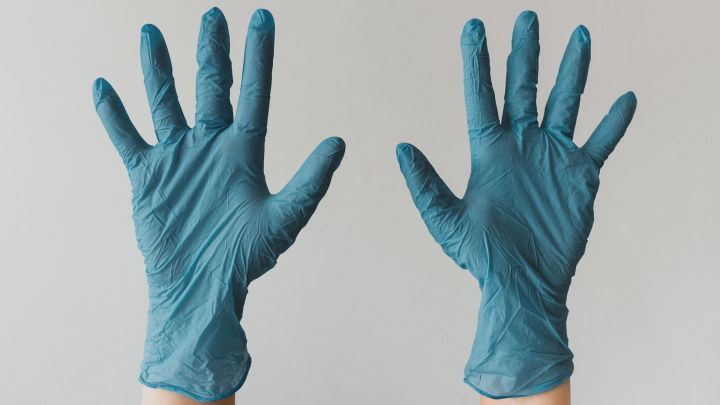 Zwei H&auml;nde in blauen Handschuhen heben sich vom neutralen Hintergrund, symbolisieren Schutz beim Einkaufen.