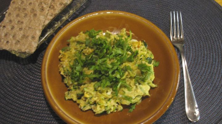 Auf einem braunen Teller liegt eine Zucchini-Eier-Pfanne, garniert mit frischen Kr&auml;utern, neben einem St&uuml;ck Brot.