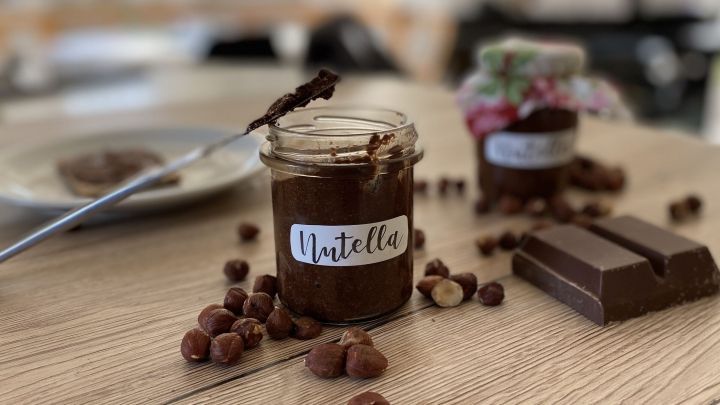Ein Glas Nutella steht auf einem Holztisch, daneben liegen Haseln&uuml;sse und Schokolade, w&auml;hrend ein L&ouml;ffel in das Glas taucht.