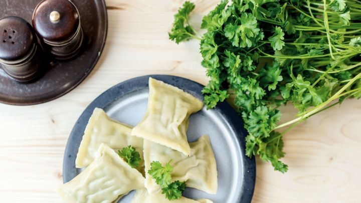 Selbstgemachte Maultaschen liegen auf einem Teller, umgeben von frischem Schnittlauch und Gew&uuml;rzgef&auml;&szlig;en auf einem Holztisch.