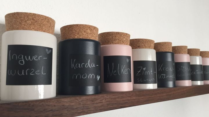 Eine Reihe von eleganten Gew&uuml;rzglasbeh&auml;ltern mit Korkdeckeln steht auf einem Holzregal an einer wei&szlig;en Wand.