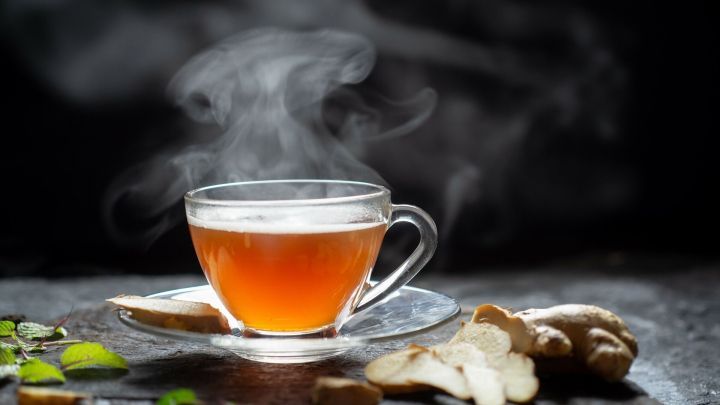 Ein dampfender Immunbooster-Tee steht auf einem Teller, umgeben von frischen Ingwerst&uuml;cken und Kr&auml;utern auf dunklem Hintergrund.