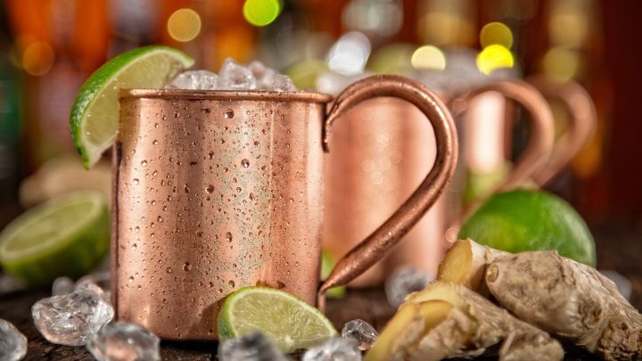 Kupferne Becher mit Eis, Limettenscheiben und Ingwer stehen auf einem Holztisch in einer bar&auml;hnlichen Umgebung.