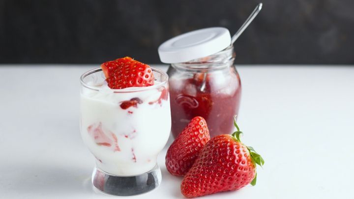 Ein Glas mit Sahne und Erdbeeren neben einem offenen Marmeladenglas, umgeben von frischen Erdbeeren auf einem hellen Tisch.