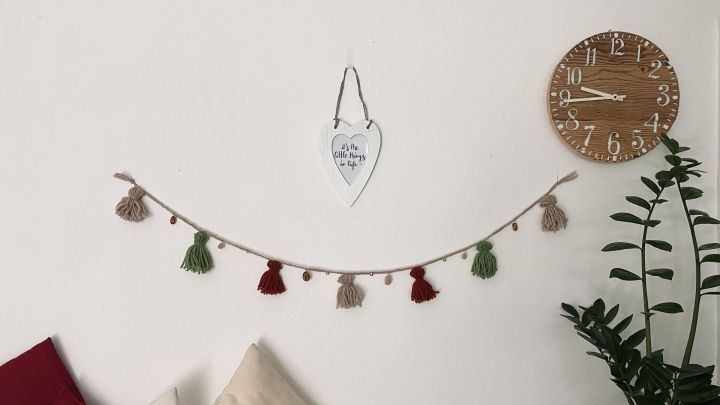 Eine Kette aus bunten Wolle-Quasten schm&uuml;ckt eine helle Wand, umgeben von Pflanzen und einem Wanduhren.