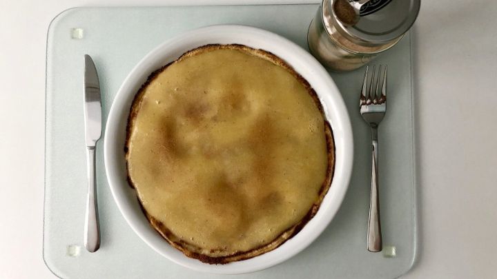 Ein runder Eierkuchen liegt in einer Sch&uuml;ssel auf einem Tisch, daneben steht ein Glas mit Strohhalm f&uuml;r zus&auml;tzlichen Genuss.