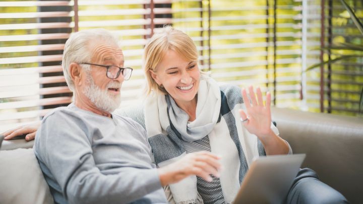 Ein &auml;lteres Paar sitzt auf einem Sofa und interagiert fr&ouml;hlich mit einem Laptop, w&auml;hrend sie sich beim Homeschooling austauschen.
