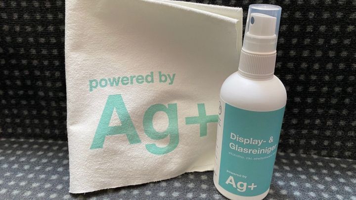 Ein Spray mit der Aufschrift &bdquo;powered by Ag+&ldquo; steht neben einem Tuch in einem modernen, strukturierten Umfeld.