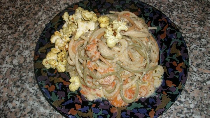 Ein Teller mit Tricolore Spaghetti, gemischt mit Karotten und K&auml;seso&szlig;e, serviert mit Thai-Curry Popcorn auf einer dunklen, gemusterten Fl&auml;che.