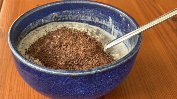 Eine blaue Sch&uuml;ssel mit M&uuml;sli steht auf einem Holztisch, w&auml;hrend ein L&ouml;ffel im M&uuml;sli steckt, das mit Schokoladenpulver bestreut ist.