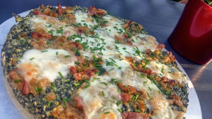 Eine k&ouml;stliche Gem&uuml;sequiche mit Giersch steht frisch auf einem Tisch im Garten, umgeben von gr&uuml;nem Laub und einem roten Becher.