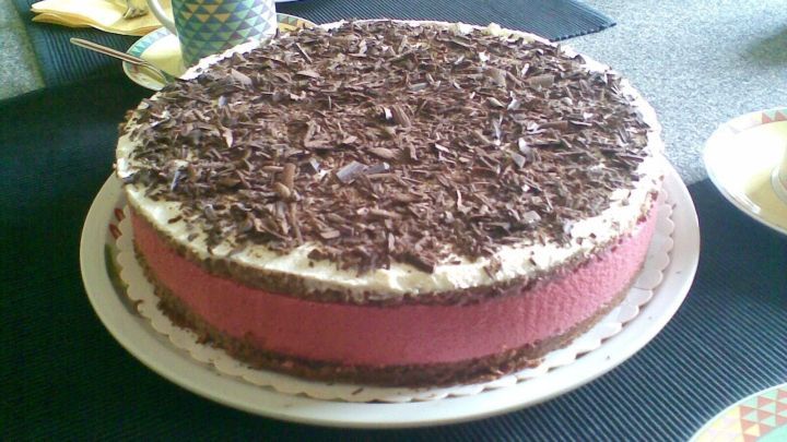 Eine Himbeertorte mit Schokobiskuitboden steht auf einem Teller, umgeben von buntem Geschirr auf einem gedeckten Tisch.