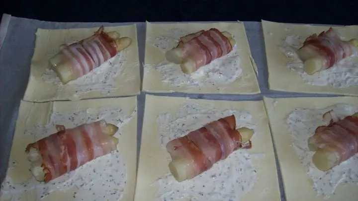 Die gleichgro&szlig; geschnittenen Spargelstangen werden nun, jeweils zwei St&uuml;cke, mit einer Scheibe Bacon umwickelt und auf die mit dem Frischk&auml;se bestrichenen Teigplatten gelegt. 