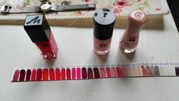 Drei Nagellackflaschen stehen neben einer Farbpalette mit verschiedenen Lackt&ouml;nen auf einem Tisch, der mit Blumenmuster dekoriert ist.