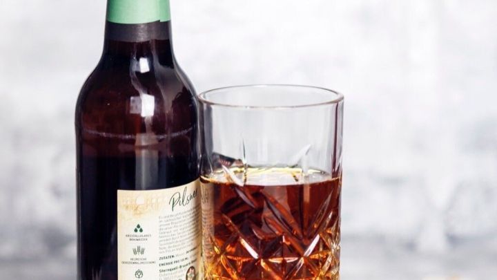 Ein Bierflasche steht neben einem Glas mit einem Bier-Korn-Cola Longdrink auf einem hellen, neutralen Untergrund.