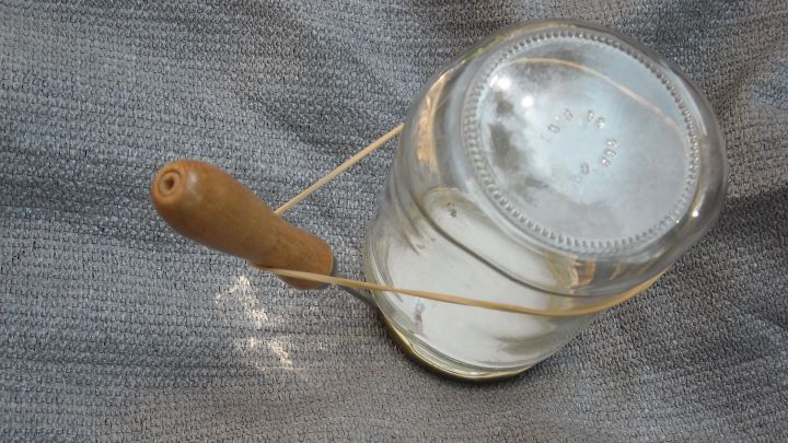 Ein Twist-Off-Glas liegt auf einem grau gemusterten Untergrund, w&auml;hrend ein Holzwerkzeug den Deckel m&uuml;helos &ouml;ffnet.