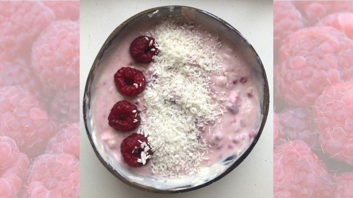 Ein cremiger Kokos-Himbeer-Joghurt in einer Sch&uuml;ssel, garniert mit Himbeeren und Kokosraspeln, vor rosa Hintergrund.