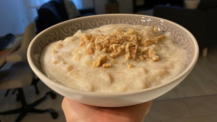 Eine handgehaltene Sch&uuml;ssel mit Fitness Grie&szlig;brei, garniert mit Honig-Apfel-Zimt-Mix, vor einem gem&uuml;tlichen Wohnzimmer.