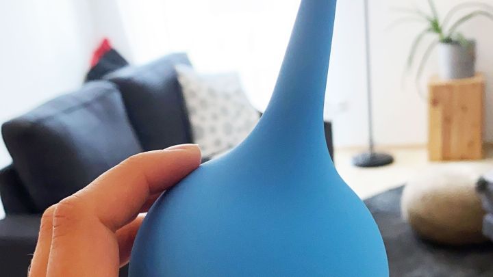 Eine Hand h&auml;lt eine blaue Ohrensp&uuml;lkanne. Sie wird verwendet, um Ohren zu reinigen. Im Hintergrund ist ein heller Wohnraum.