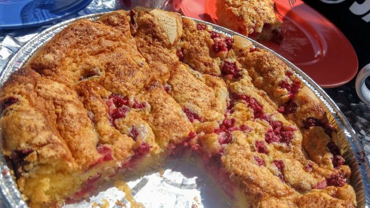 Ein k&ouml;stlicher Himbeerkuchen mit einer goldenen, knusprigen Kruste wird auf einem Tisch serviert, umgeben von Tellern und einem Glas mit frischen Beeren.