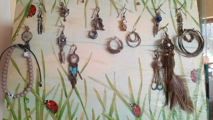 Ein Ohrringhalter zeigt verschiedene Ohrringe, die an einer bemalten Holzplatte im Blumenmotiv hangen, umgeben von einer wohnlichen Umgebung.