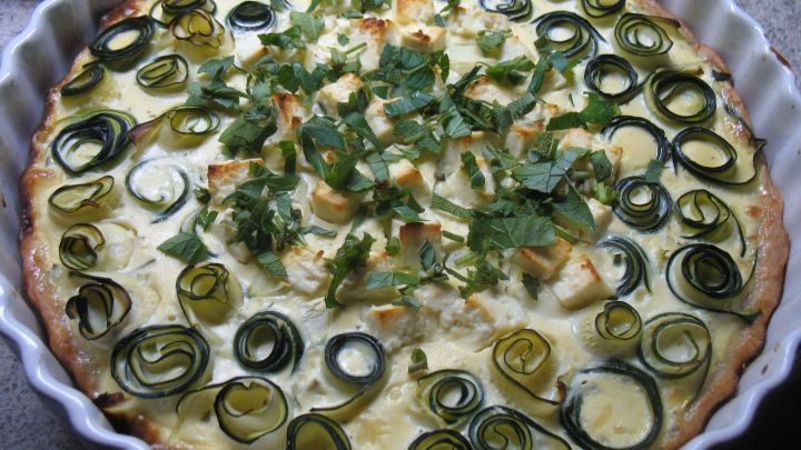 Eine frisch gebackene Zucchini-Tarte mit K&auml;se und Kr&auml;utern liegt in einer runden Form, dekoriert mit gr&uuml;nen Zucchinischeiben.