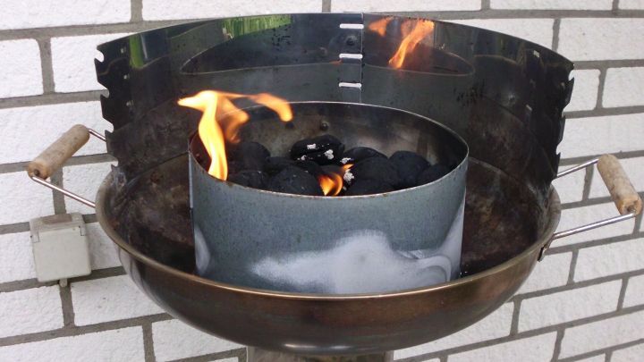 Eine Gartengrill mit brennenden Kohlen in einer zentralen Metallpfanne steht auf einem Steinboden vor einer Ziegelwand.