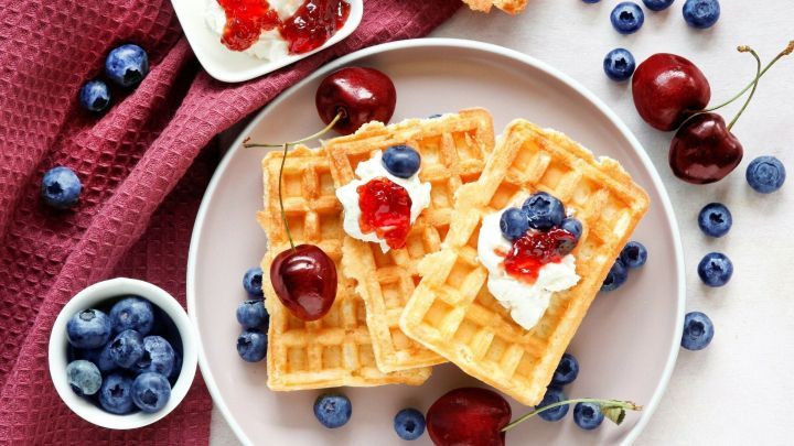 Waffeln auf einem Teller liegen dekoriert mit Sahne, Beeren und Gelee, umgeben von frischen Fr&uuml;chten und einem Tuch.