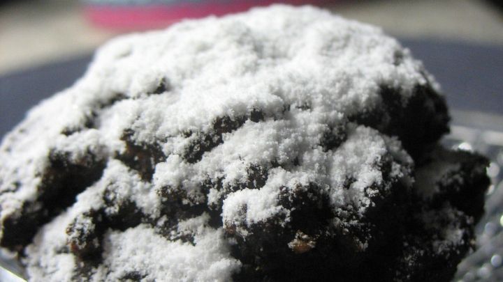 Ein Schoko-Aprikosen-Muffin sitzt auf einer gl&auml;sernen Oberfl&auml;che, pudriger Zucker bedeckt seine Oberseite, umgeben von sanfter Beleuchtung.