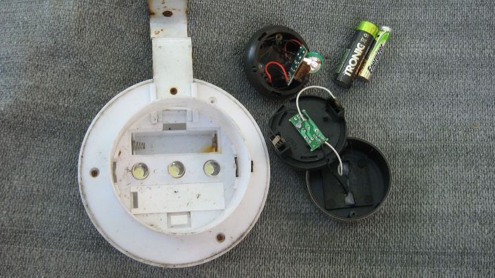 Eine Solargartenlampe liegt zerlegt auf einem grauen Untergrund, mit Batterien und Einzelteilen, um sie wieder zum Leuchten zu bringen.
