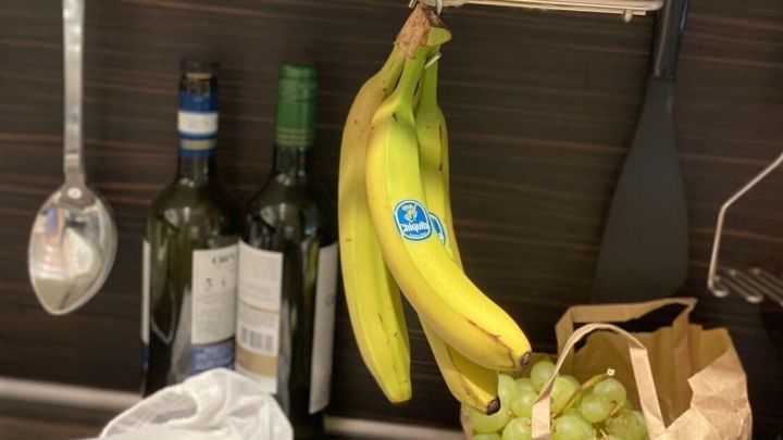 Gelbe Bananen h&auml;ngen in einer K&uuml;che &uuml;ber einem Regal, w&auml;hrend Trauben in einer Papiert&uuml;te neben einer Flasche stehen.