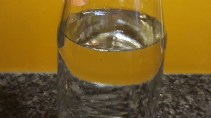 Ein Glas mit Wasser steht auf einer dunklen Oberfl&auml;che, w&auml;hrend es von einem hellen, gelben Hintergrund umgeben ist.