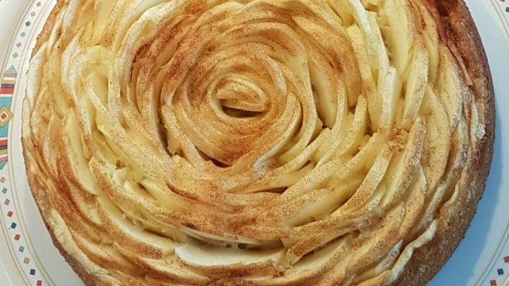 Eine kunstvoll dekorierte Apfelkuchentorte, geformt wie eine gro&szlig;e Rosenbl&uuml;te, ruht auf einem bunten Teller.