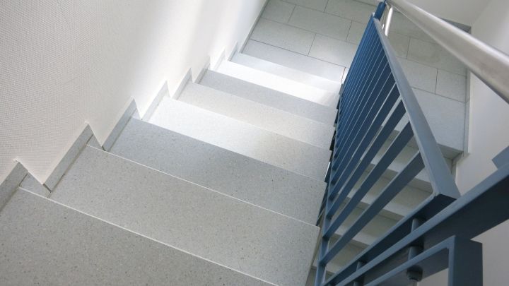 Kellertreppe mit einer rutschfesten Oberfl&auml;che und blauen Handlauf, die in einem hellen, modernen Raum f&uuml;hrt.
