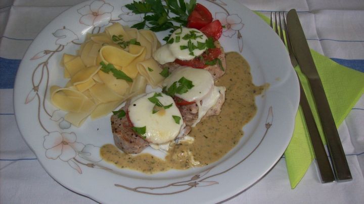 Saftige, gratinierte Schweinemedaillons liegen auf einem Teller, umgeben von Nudeln und Basilikumso&szlig;e, serviert mit Tomate.